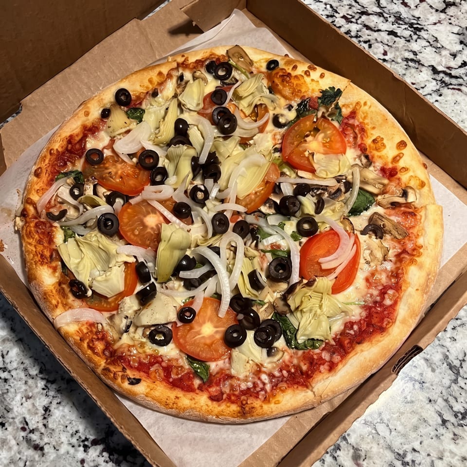 A whole pizza in a cardboard box, topped with tomatoes, olives, onions, and artichoke hearts.