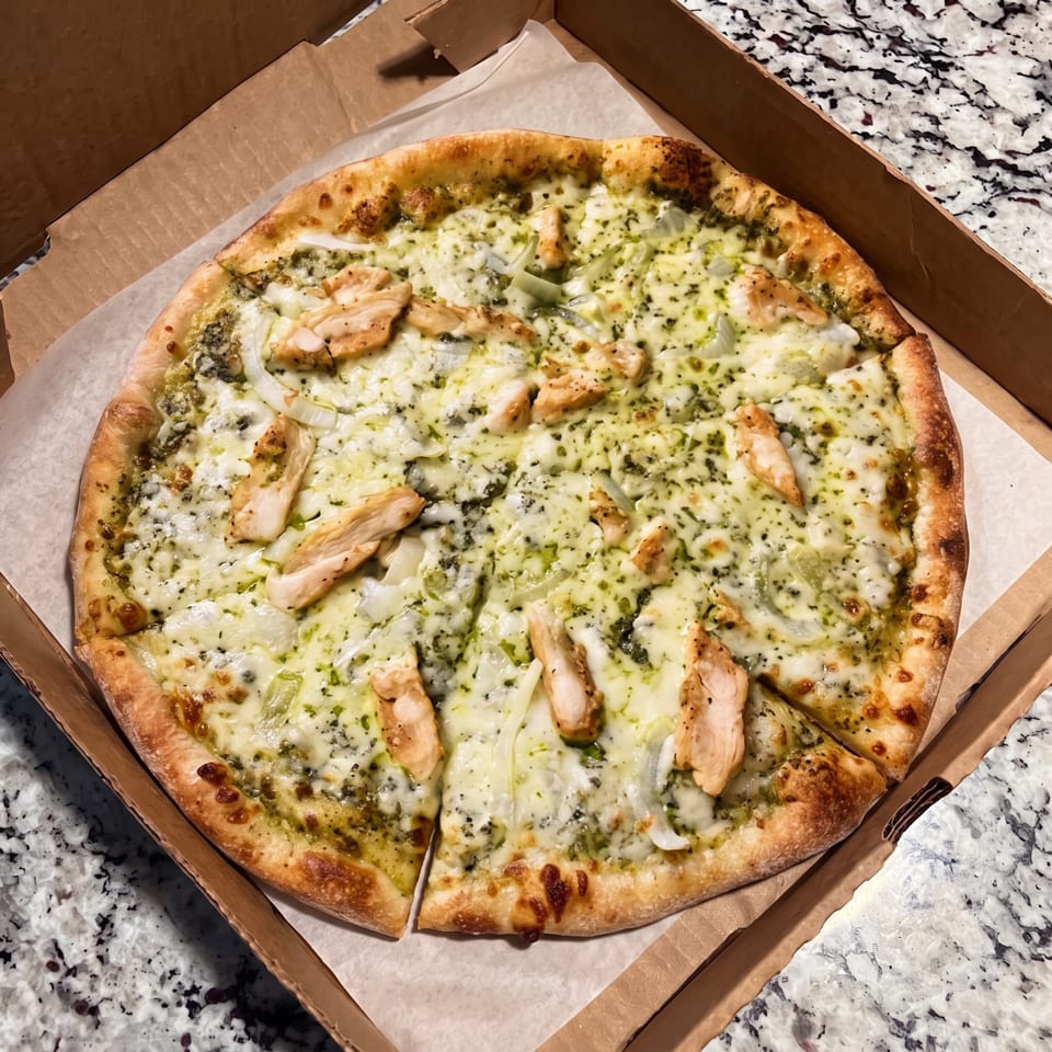 A whole pesto chicken pizza with white cheese and onions in a cardboard delivery box.